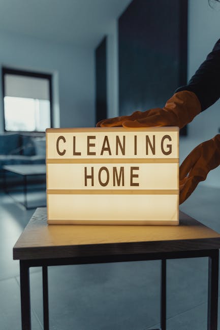 A person wearing a brown glove is adjusting or placing a lightbox sign on a wooden surface, with the illuminated sign displaying the words 'CLEANING HOME' in bold capital letters. The room features a 